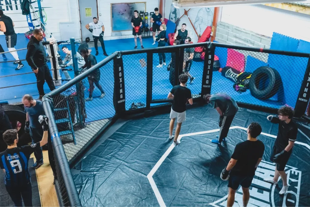 Groupe de personnes s'entraînant au MMA dans un ring de boxe spécialisé. En arrière plan d'autres gens évoluent dans la salle.