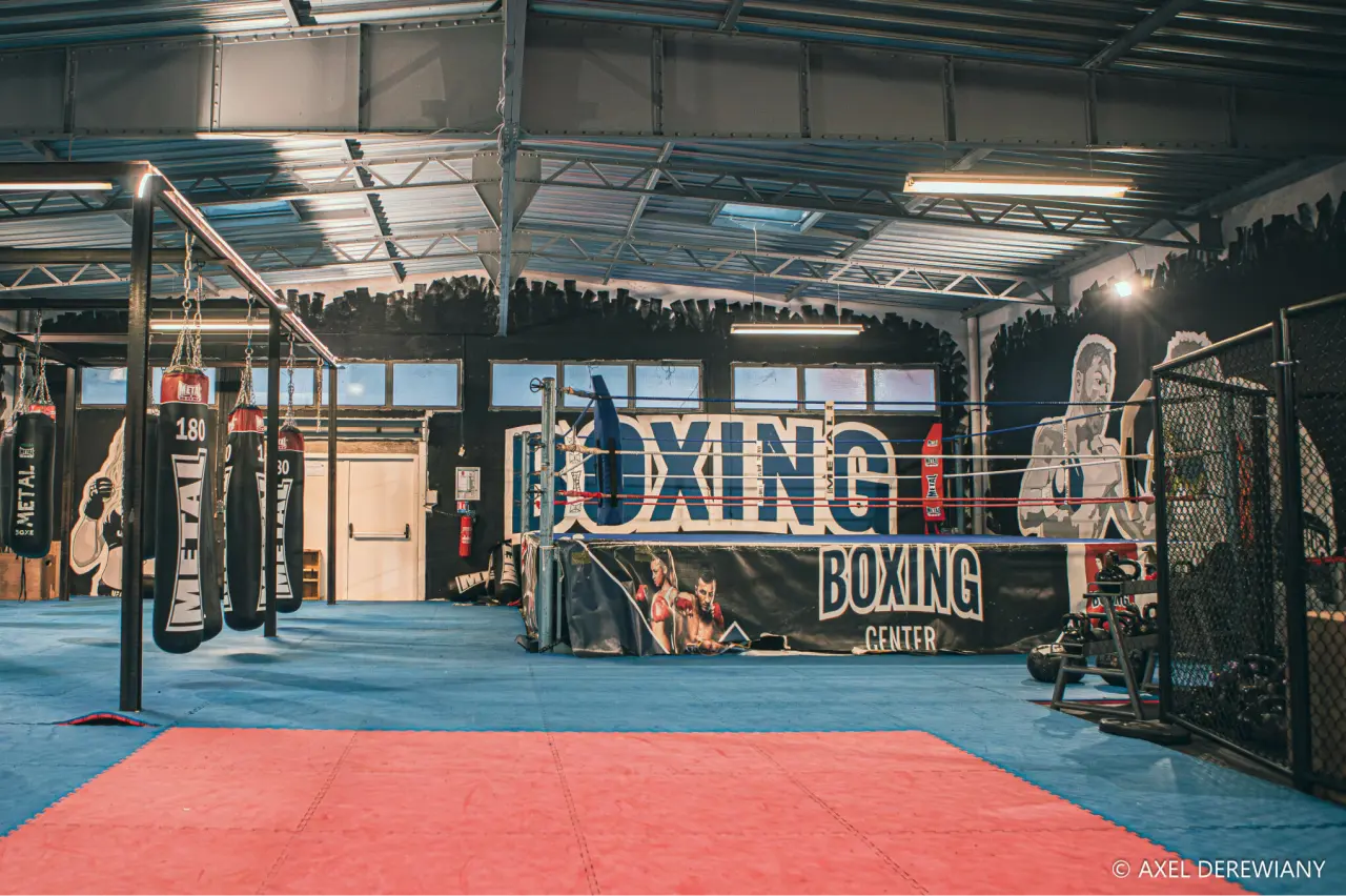 Une salle de boxe avec un ring dans le fond à droite, des sacs de frappes à gauche.