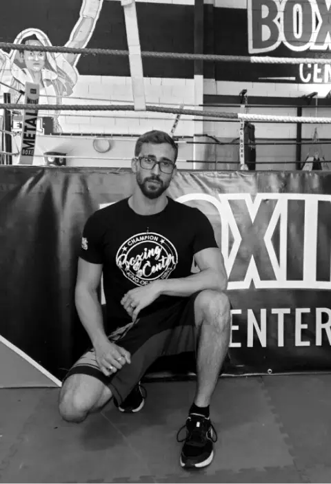 Un jeune homme avec une barde portant des lunettes. il est accroupi devant un ring de boxe.