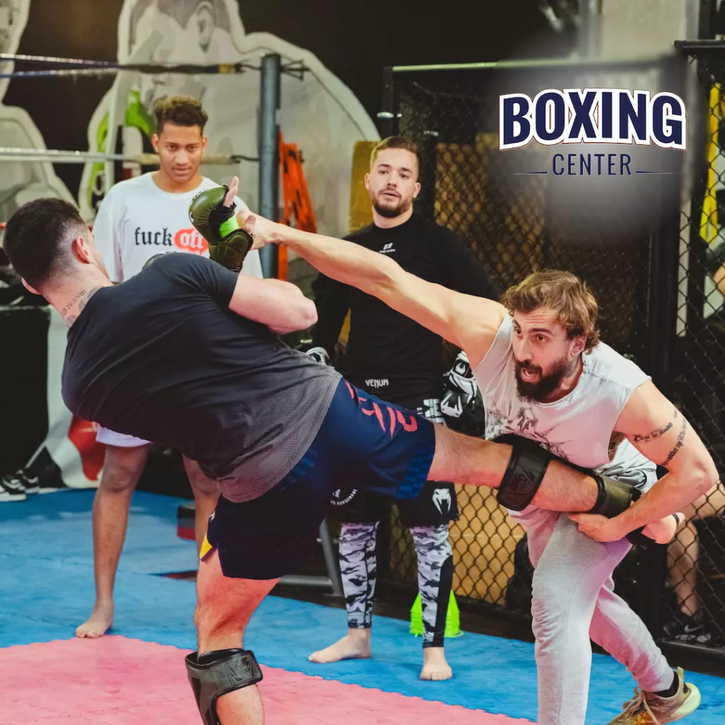 Deux hommes pratiquent les arts martiaux dans une salle de sport, l'un d'eux donne un coup de pied. Trois autres observent attentivement. Le logo « Boxing Center » est visible. Ambiance intense et concentrée.